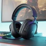 Headset gamer preto com iluminação RGB sobre uma mesa com teclado mecânico, mouse e monitor de computador ao fundo.