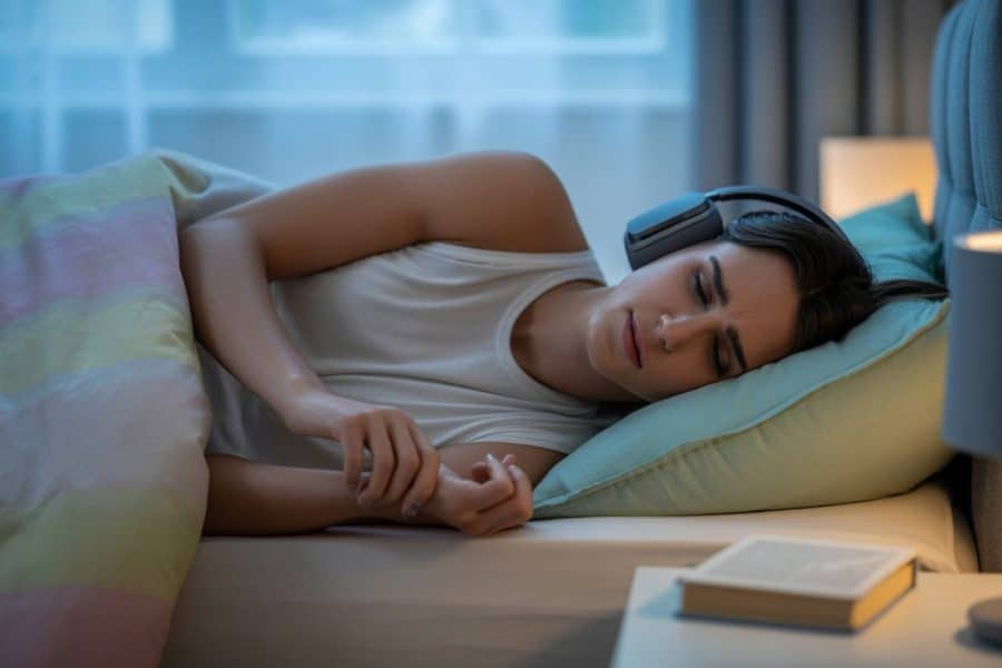 Pessoa deitada na cama usando fones de ouvido, com expressão desconfortável, em um quarto iluminado naturalmente.
