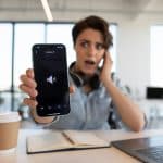 Pessoa sentada em uma mesa de escritório olhando surpresa para o celular com o volume baixo.
