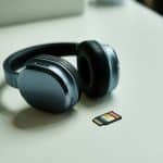 Fone de ouvido moderno preto com acabamento metálico ao lado de um cartão de memória microSD sobre uma superfície branca.