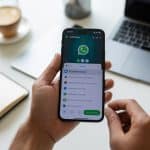 Pessoa segurando um smartphone com o aplicativo WhatsApp aberto, ajustando configurações em um ambiente de trabalho organizado.