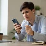 Pessoa segurando um celular e um fone com fio, olhando para o conector com expressão de dúvida em uma mesa de escritório em casa.