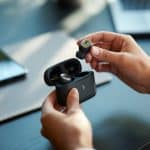 Mãos segurando fones de ouvido Bluetooth e colocando-os em um estojo de carregamento aberto sobre uma mesa.