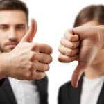 Duas mãos, uma fazendo sinal de positivo com o polegar para cima e outra fazendo sinal de negativo com o polegar para baixo, em fundo neutro e desfocado.