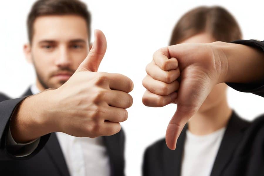 Duas mãos, uma fazendo sinal de positivo com o polegar para cima e outra fazendo sinal de negativo com o polegar para baixo, em fundo neutro e desfocado.