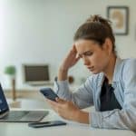 Pessoa jovem sentada à mesa em escritório moderno, olhando preocupada para a tela do celular ou laptop.