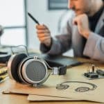 Fones de ouvido clássicos sobre uma mesa de madeira com esboços e ferramentas ao redor, e uma pessoa ao fundo examinando um protótipo.