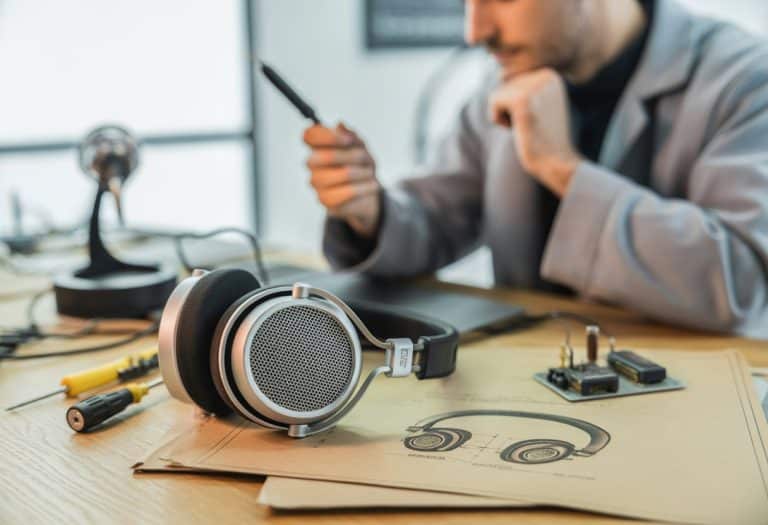 Fones de ouvido clássicos sobre uma mesa de madeira com esboços e ferramentas ao redor, e uma pessoa ao fundo examinando um protótipo.