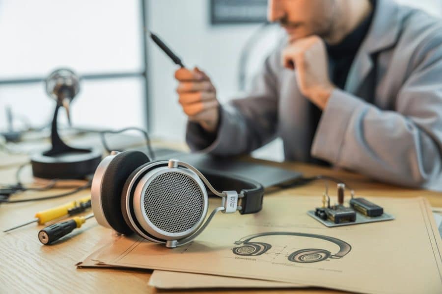 Fones de ouvido clássicos sobre uma mesa de madeira com esboços e ferramentas ao redor, e uma pessoa ao fundo examinando um protótipo.