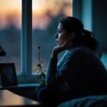 Pessoa sentada sozinha em um quarto com luz suave, olhando pensativa pela janela ao entardecer.