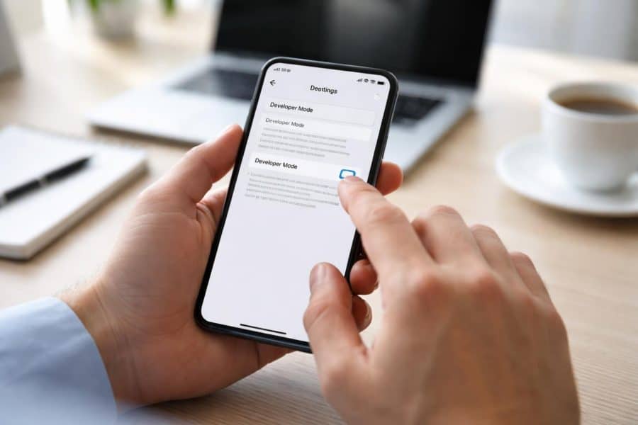 Pessoa segurando um smartphone com menus de configurações, em uma mesa com laptop e café ao fundo.