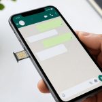 Uma mão segurando um celular com o chip parcialmente removido, mostrando a tela do WhatsApp aberta.