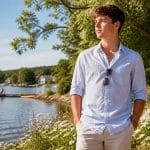 Um jovem em um parque ensolarado de verão, olhando pensativo para o horizonte próximo a um lago tranquilo.